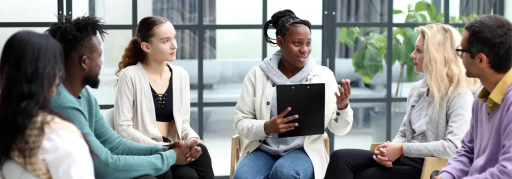 Group of five colleagues sitting in a circle in a modern office, smiling and engaged in conversation; a woman in the middle holds a tablet.