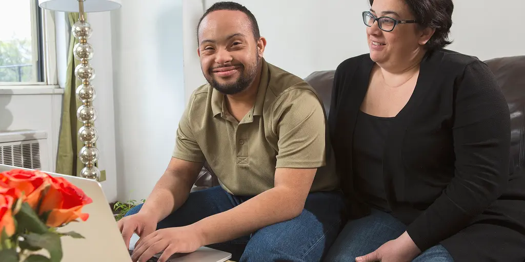 Smiling man sitting on a couch using a laptop, with a woman wearing glasses sitting beside him and smiling.