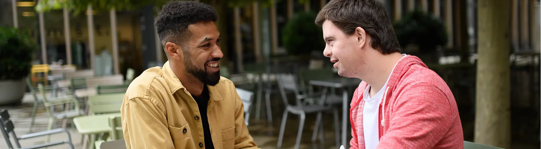 Two men sitting at an outdoor café engaged in conversation; one wears a tan jacket and the other a red cardigan, both smiling.