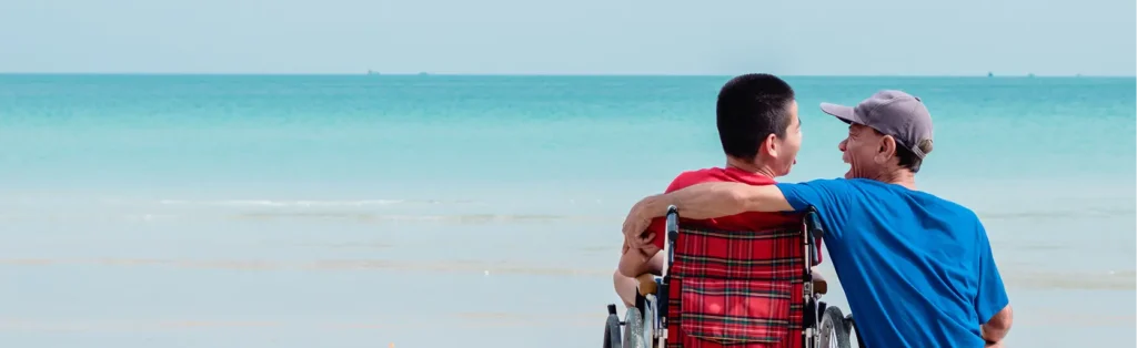 Two friends at the beach with their arms around each other; one sits in a wheelchair while the other stands beside him, both looking toward the ocean.