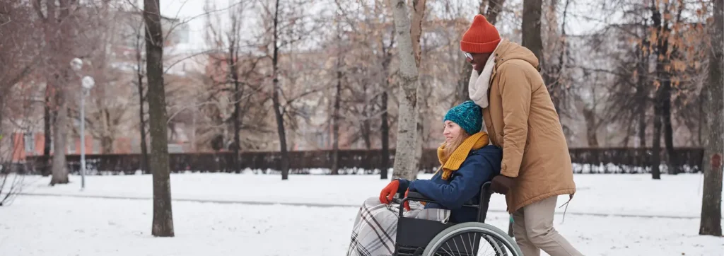 Care partner wearing winter clothing pushes a smiling person in a wheelchair through a snow-covered park.