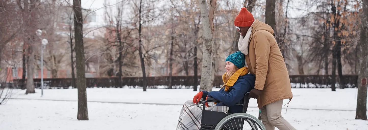 Care partner wearing winter clothing pushes a smiling person in a wheelchair through a snow-covered park.