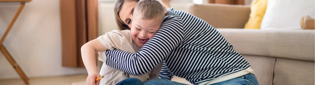 Smiling woman hugging a laughing boy in a striped sweater as they sit on a sofa at home.