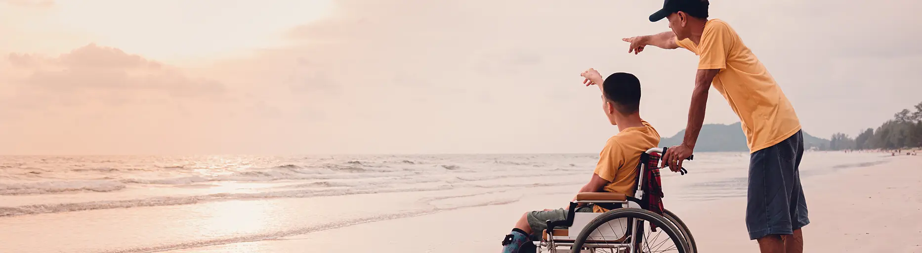Care partner in a yellow shirt pushes a boy in a wheelchair along the beach at sunset, pointing toward the waves.