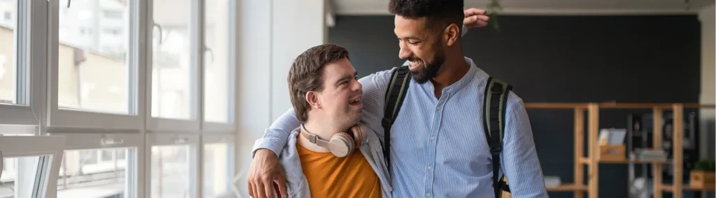 Two young men stand arm-in-arm in a hallway with large windows; one wears headphones around his neck and a yellow sweater, and the other carries a backpack; both are smiling at each other.