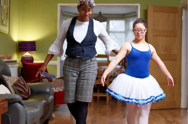 AdobeStock_481488935 1 Dance instructor wearing a vest and hat guides a young woman in a blue leotard and white tutu through ballet steps in a living room.
