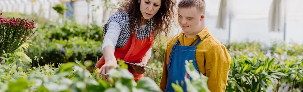 Woman in a red apron shows a young person with a short haircut and yellow apron how to tend plants in a greenhouse.