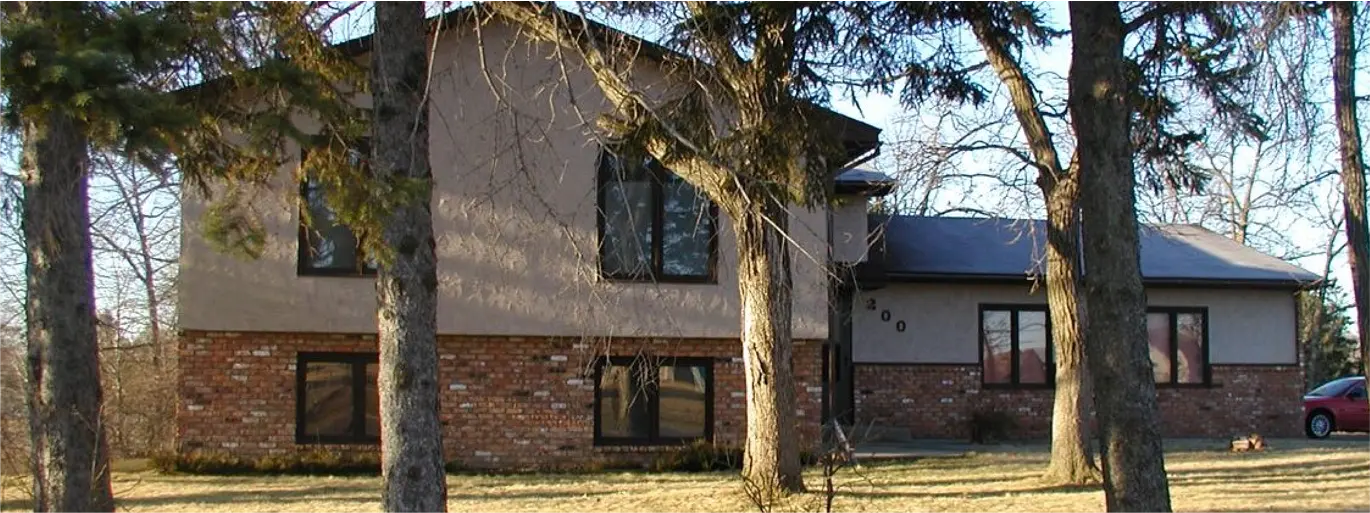 Two-story house with a brick lower level and beige upper level, partly obscured by tall pine trees in the front yard