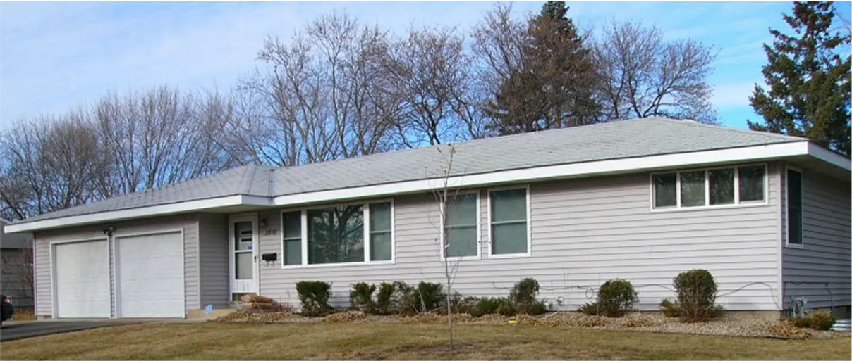 Single-story gray ranch house with an attached garage, small shrubs in front, and leafless trees in the background