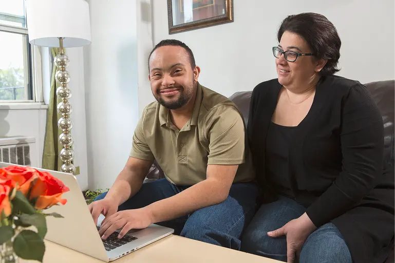 Young man smiling and using a computer with a woman sitting next to him Smiling man sitting at a table using a laptop, with a woman wearing glasses sitting beside him and smiling.