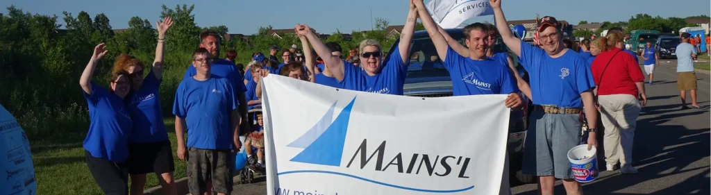 Group of people wearing blue Mains’l shirts standing outside holding a large Mains’l banner, cheering and raising their hands.