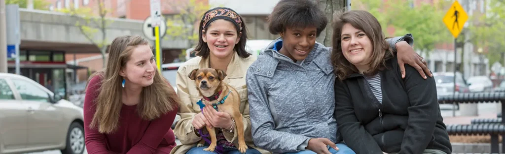Four young women sitting together outdoors on a bench, smiling; one of them holds a tan dog on her lap.