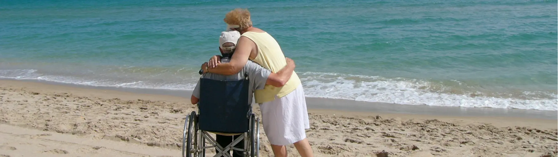 Care partner hugging an elderly person in a wheelchair on a sandy beach by the sea.