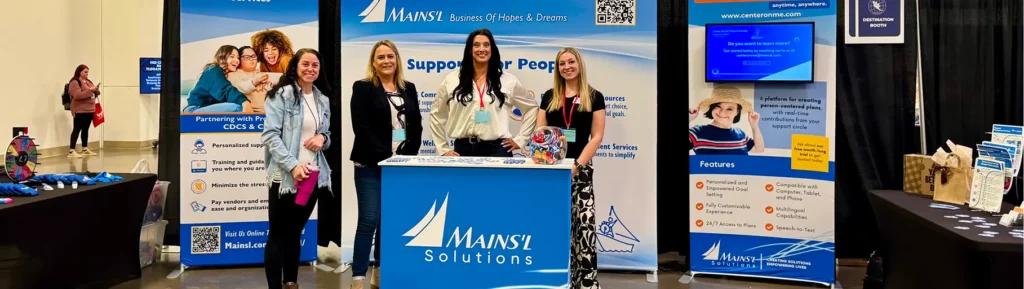 A Mains’l Solutions booth at an event with four women standing behind the table amid banners and displays.