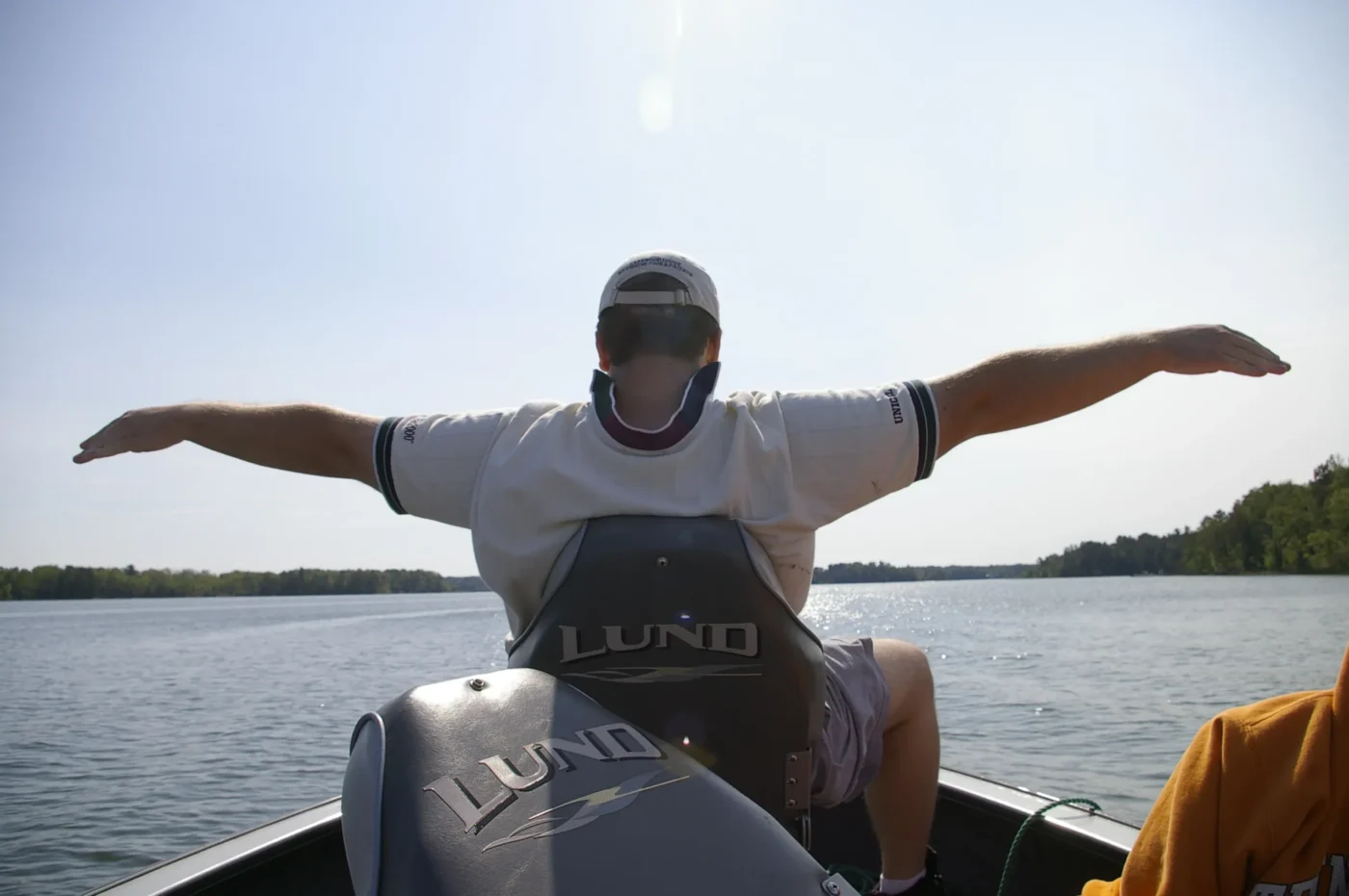 Person sitting at the front of a boat with arms outstretched, wearing a cap and face covering, with water and a forested shoreline in the background.