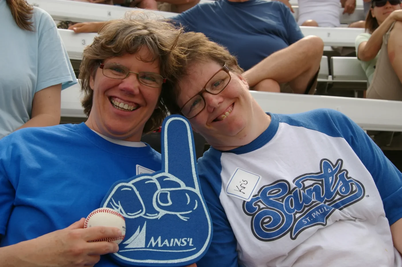 Two people smile for the camera at a St. Paul Saints baseball game wearing team gear, with a foam finger and scoreboard in the background.