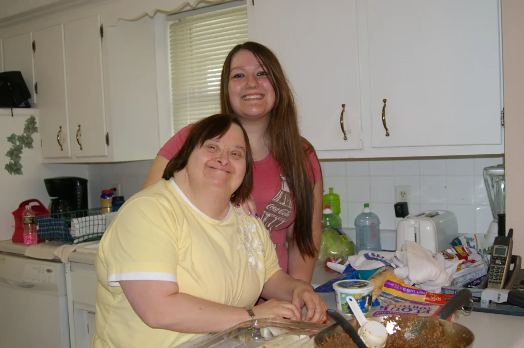 Two people in a kitchen smiling together while preparing food; one person sits at a table and the other stands behind with arms around them.