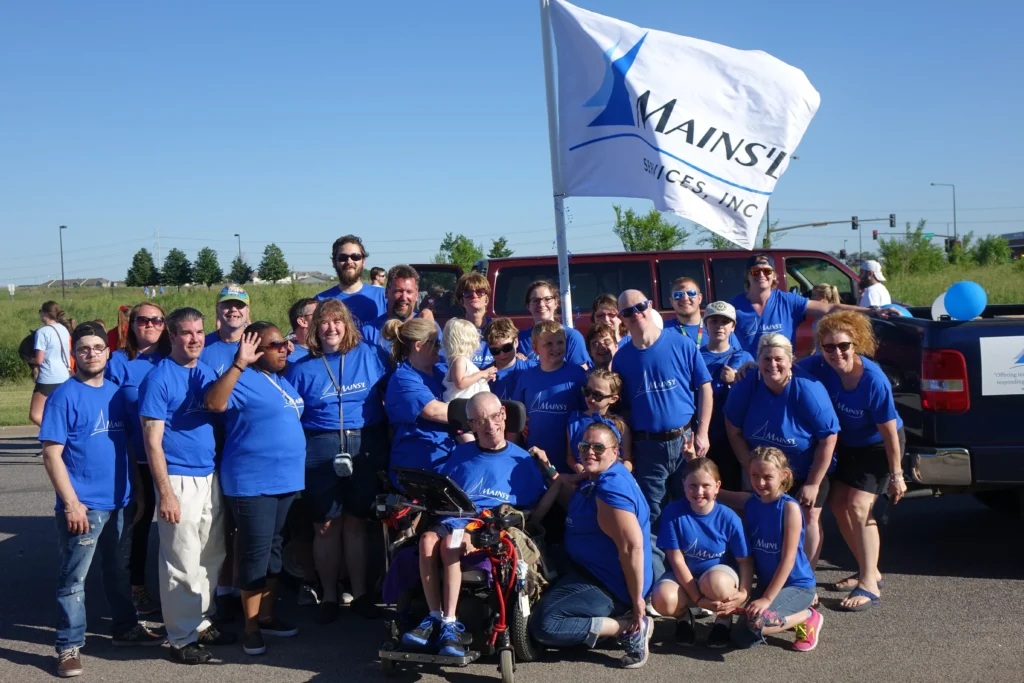 Large group of people wearing matching blue shirts posing outdoors with a company flag and a vehicle; some standing, some crouching, and one person in a wheelchair.