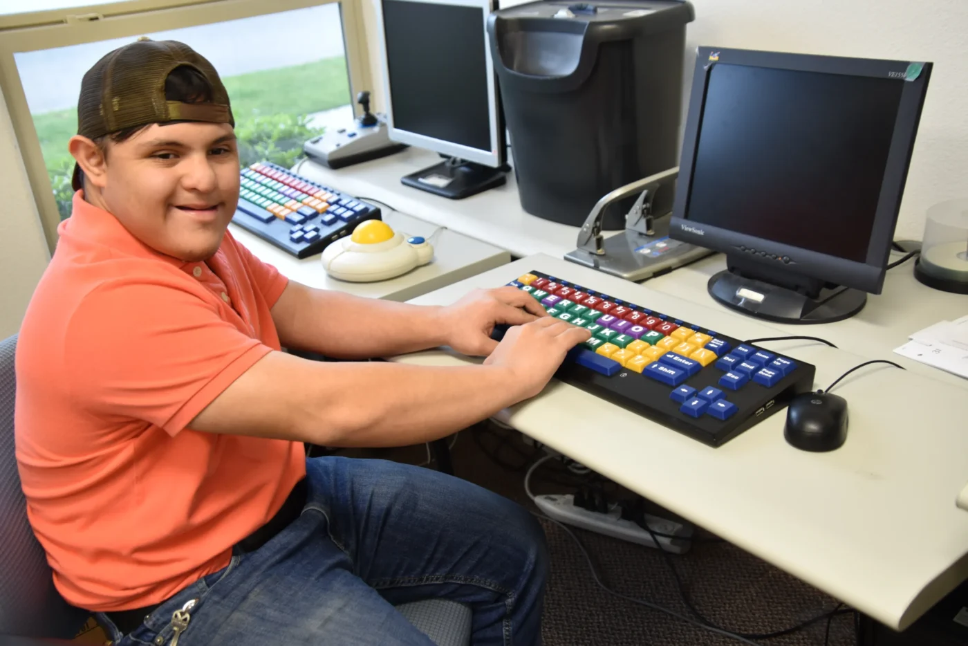 Person wearing a cap and orange shirt sitting at a desk using a computer with a large colorful keyboard and trackball.