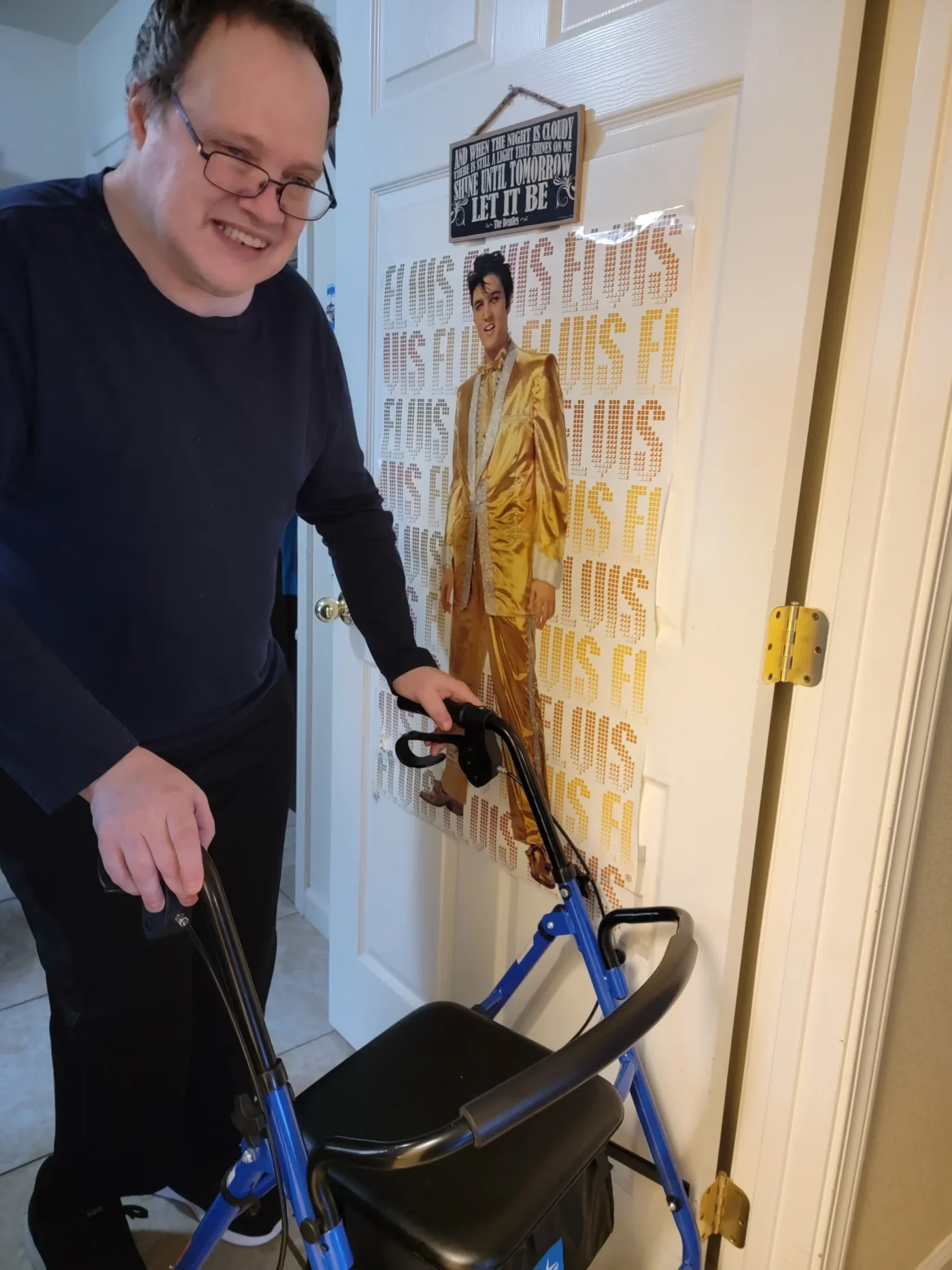 Man smiling while using a blue walker in his home hallway, standing near a door decorated with an Elvis Presley poster.