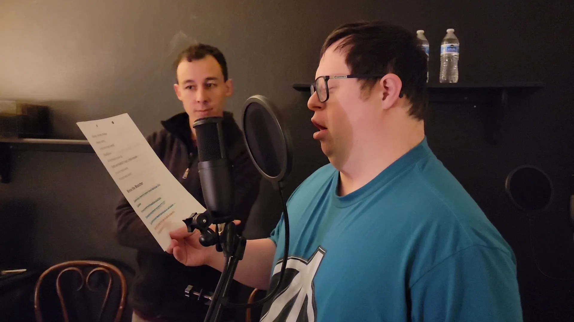20250101-man-rehearses-in-audio-booth-for-play-mainsl-california Man wearing glasses and a teal shirt rehearsing at a microphone in an audio booth, holding a script while another person stands behind him.