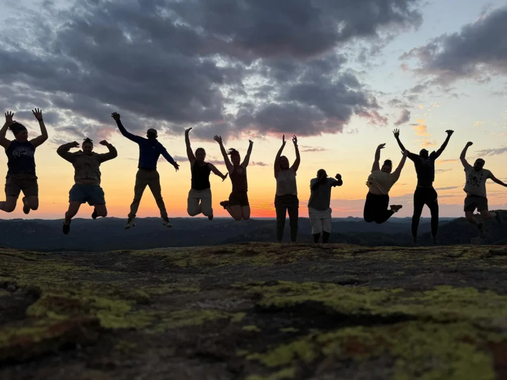 Silhouetted group of people jumping and raising their arms on a rocky outcrop at sunset, with dramatic clouds and a colorful sky in the background.