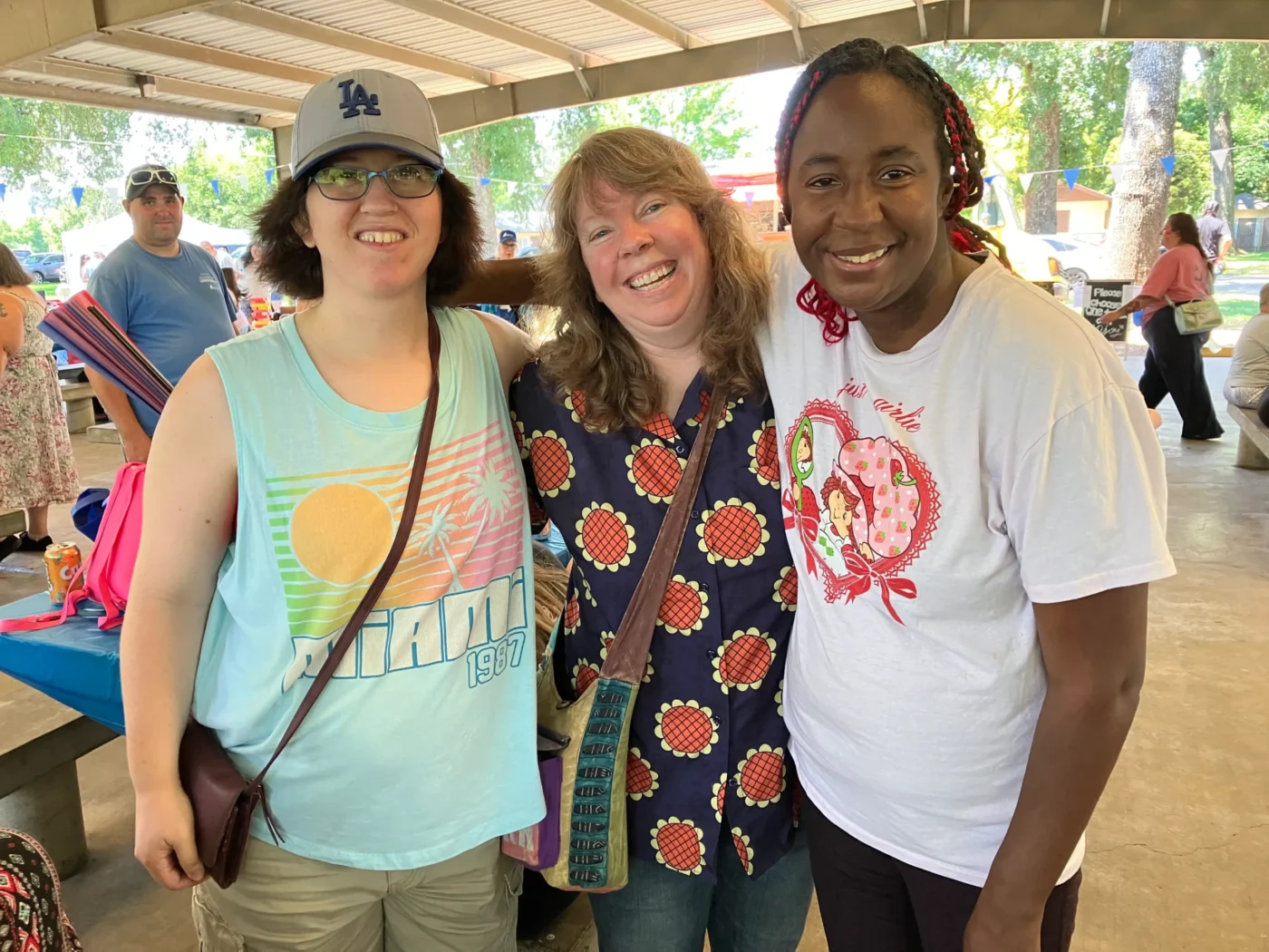 Three women standing close together and smiling at an outdoor picnic under a pavilion; one wears a baseball cap and Miami shirt, while the others hug her and smile.