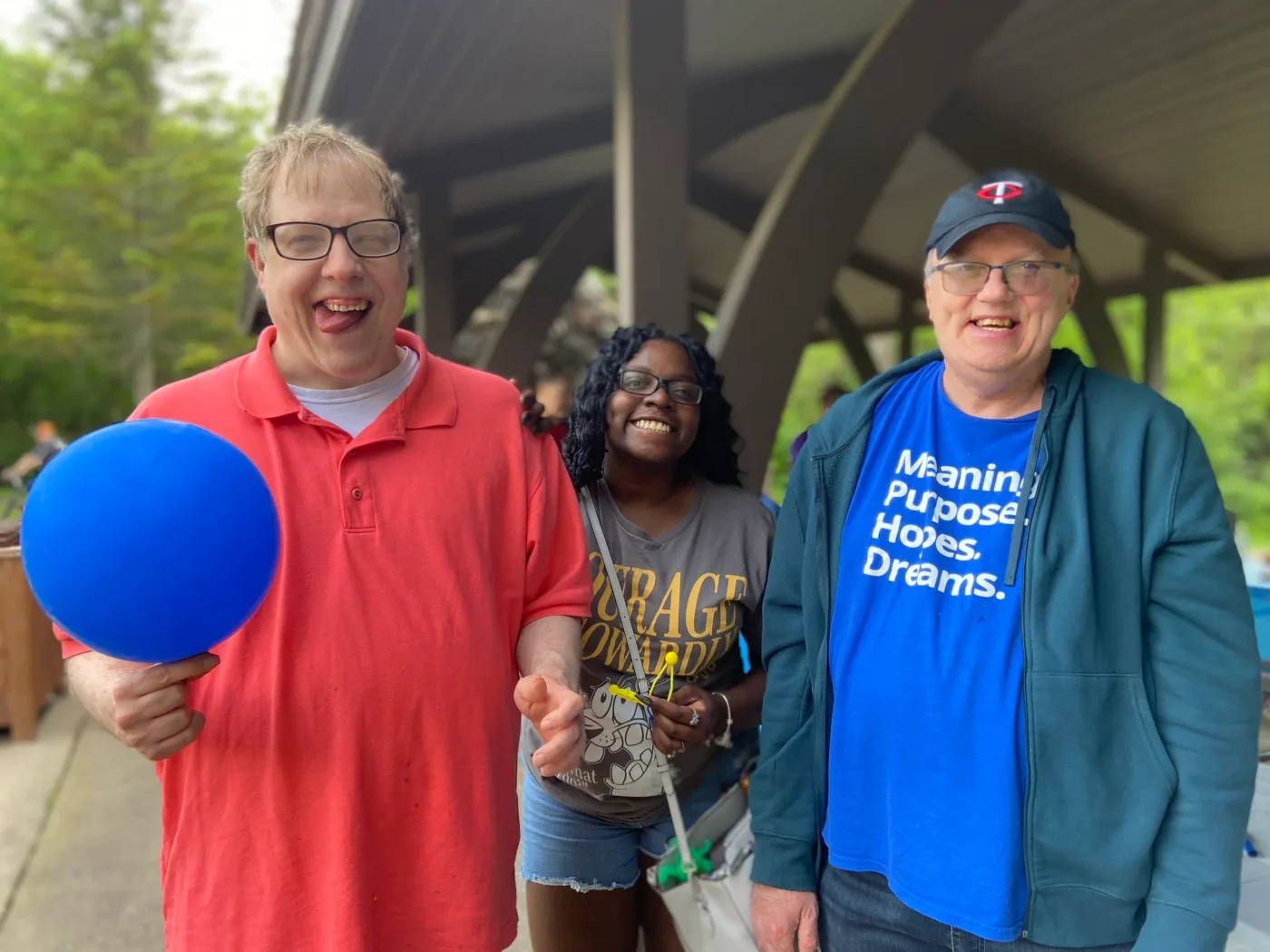 Three people smiling at an outdoor picnic; a man in a coral shirt holds a blue balloon, a woman stands in the middle, and another man in a blue jacket and baseball cap stands beside them.