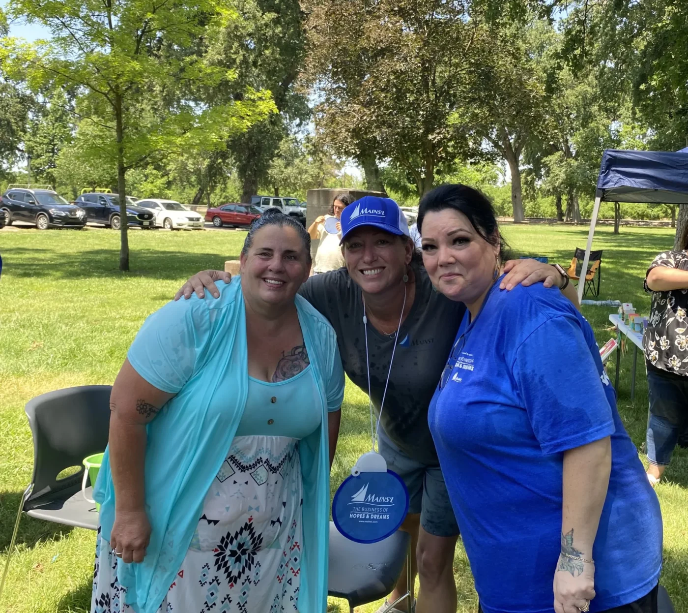 Three women standing together outdoors at a park, smiling at the camera during a community gathering.