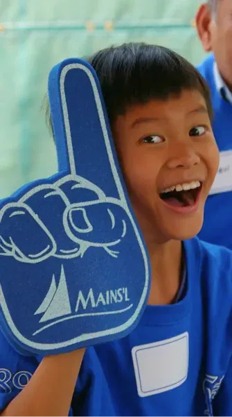 Smiling boy wearing a blue shirt holding up a large Mains’l branded number one foam finger.