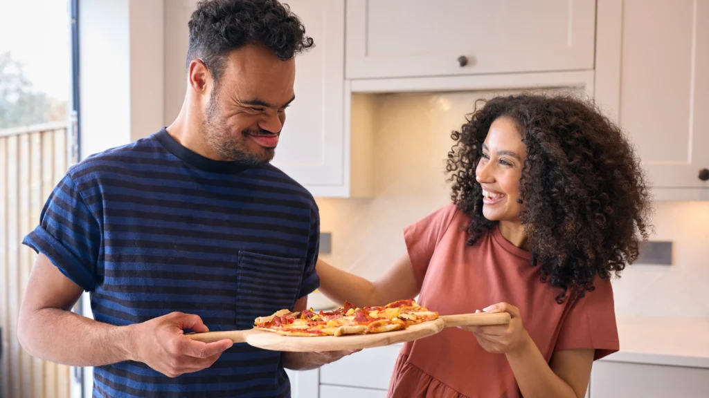 A man with Down Syndrome and a woman prepare to eat a pizza in a home kitchen.