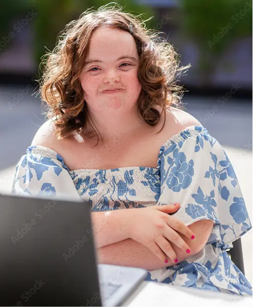 Young woman with curly hair sitting outdoors in front of a laptop, smiling gently at the camera.