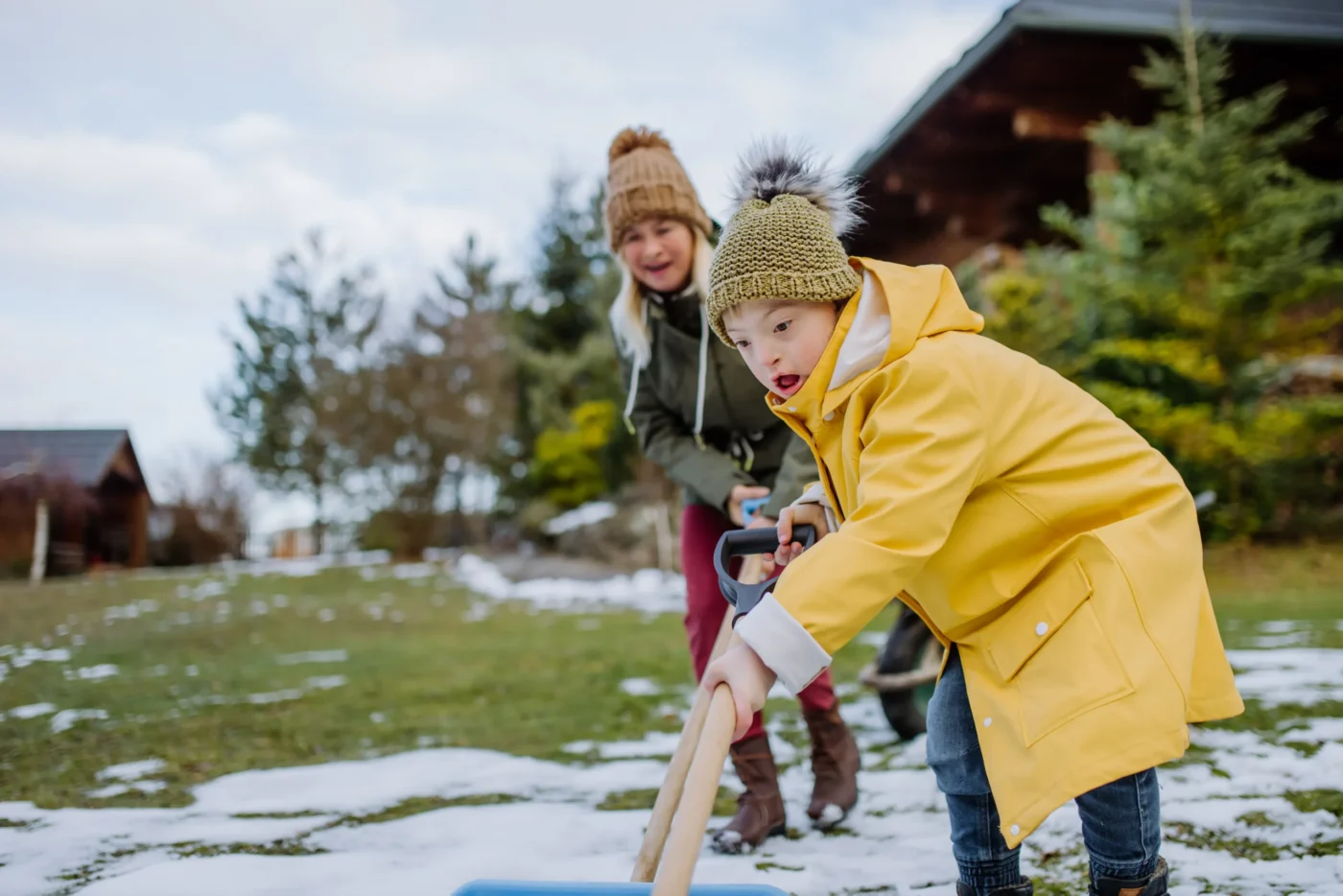adult-and-child-with-down-syndrome-shovel-snow-outside Child wearing a yellow coat shoveling snow outdoors with an adult in winter clothing behind them, with houses and trees in the background.