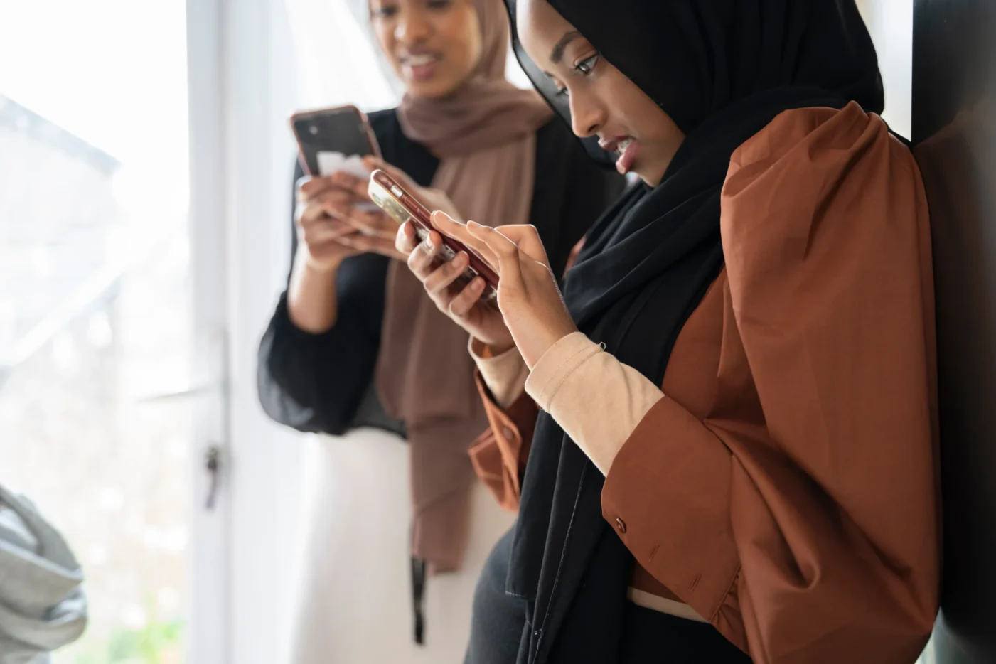 Woman wearing a hijab intently using a smartphone while another woman in the background also looks at her phone, standing in a bright room.