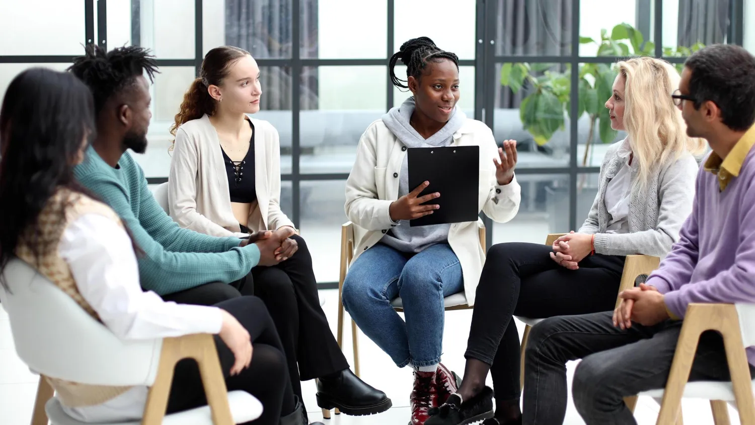 Group of adults seated in a circle having a discussion in a bright room, with one person gesturing while others listen attentively.