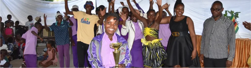 Group of people celebrating indoors, with one person in front wearing a purple outfit and holding a trophy while others stand behind raising their arms.