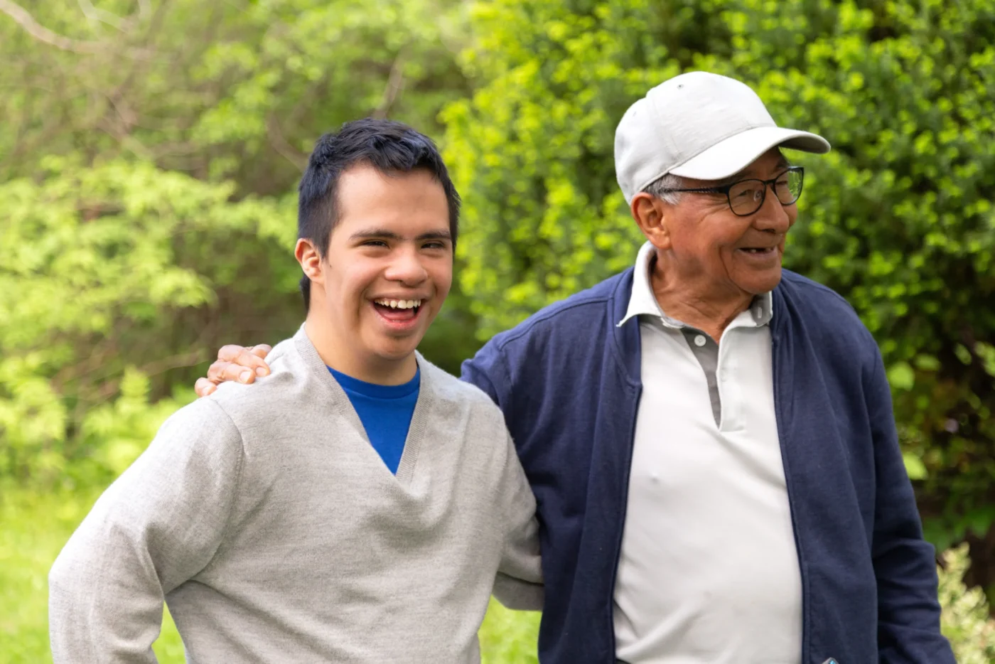 Older man wearing a hat and glasses walking outdoors with a younger man, both smiling with an arm around each other's shoulders.