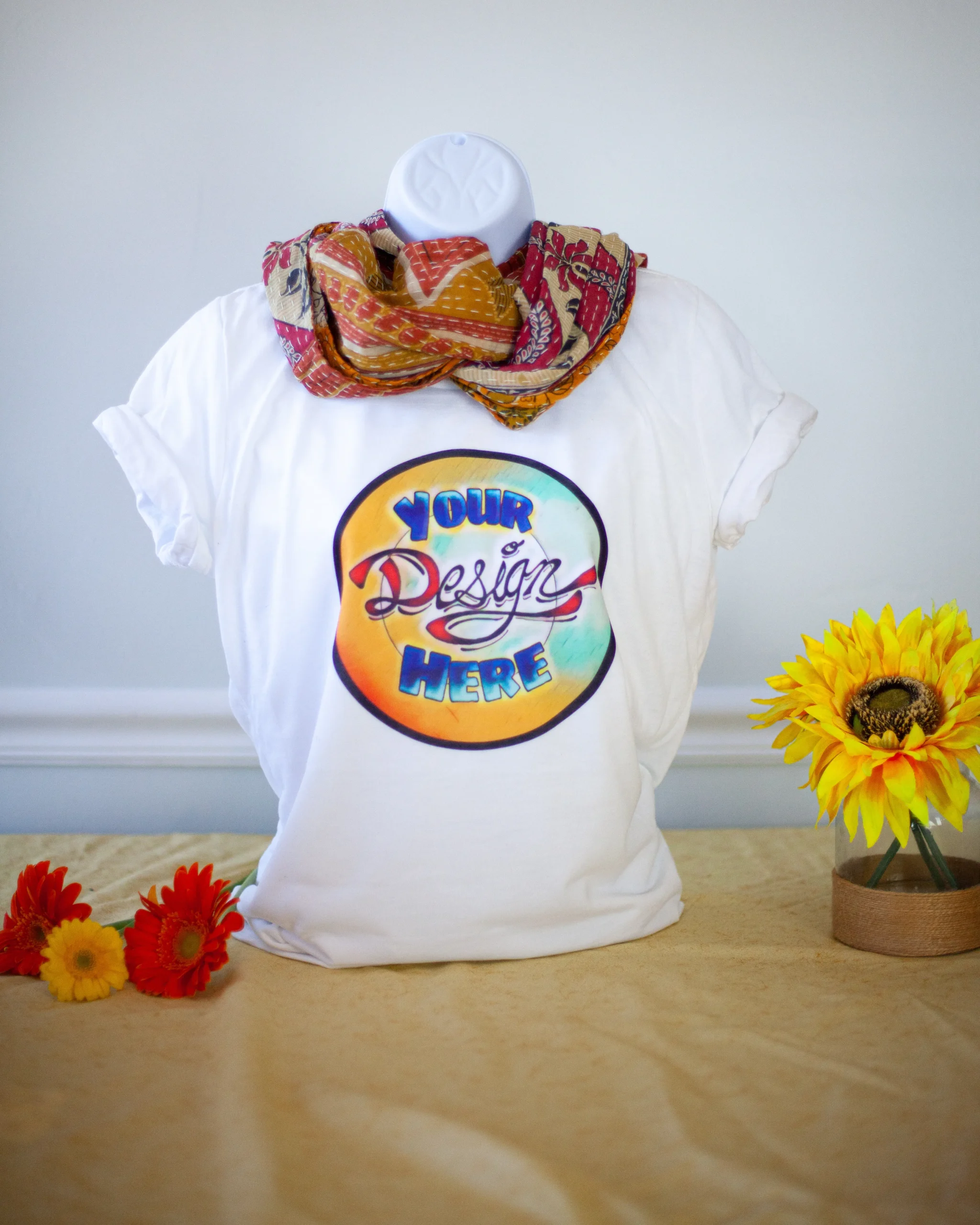 White T-shirt displayed on a mannequin with a colorful "Your Design Here" circle graphic, accessorized with a patterned scarf and surrounded by flowers on a table.