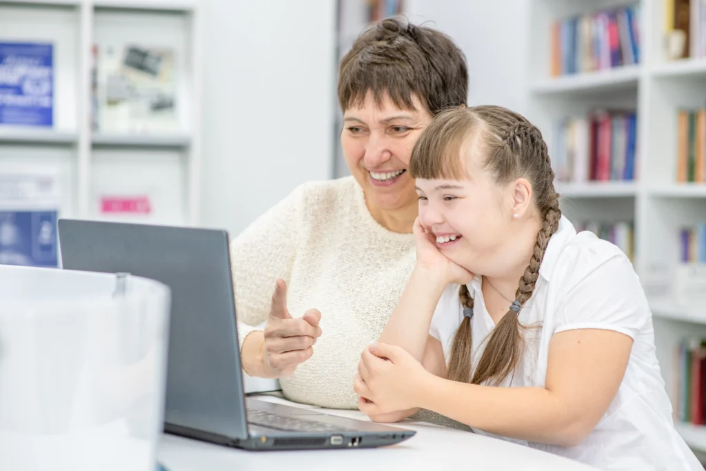 Older woman sitting with a younger woman with Down syndrome, both smiling as they look at a laptop together