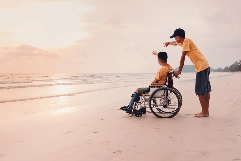 Person in a wheelchair with a friend at the beach during sunset, raising an arm as the friend stands behind them with ocean waves in view