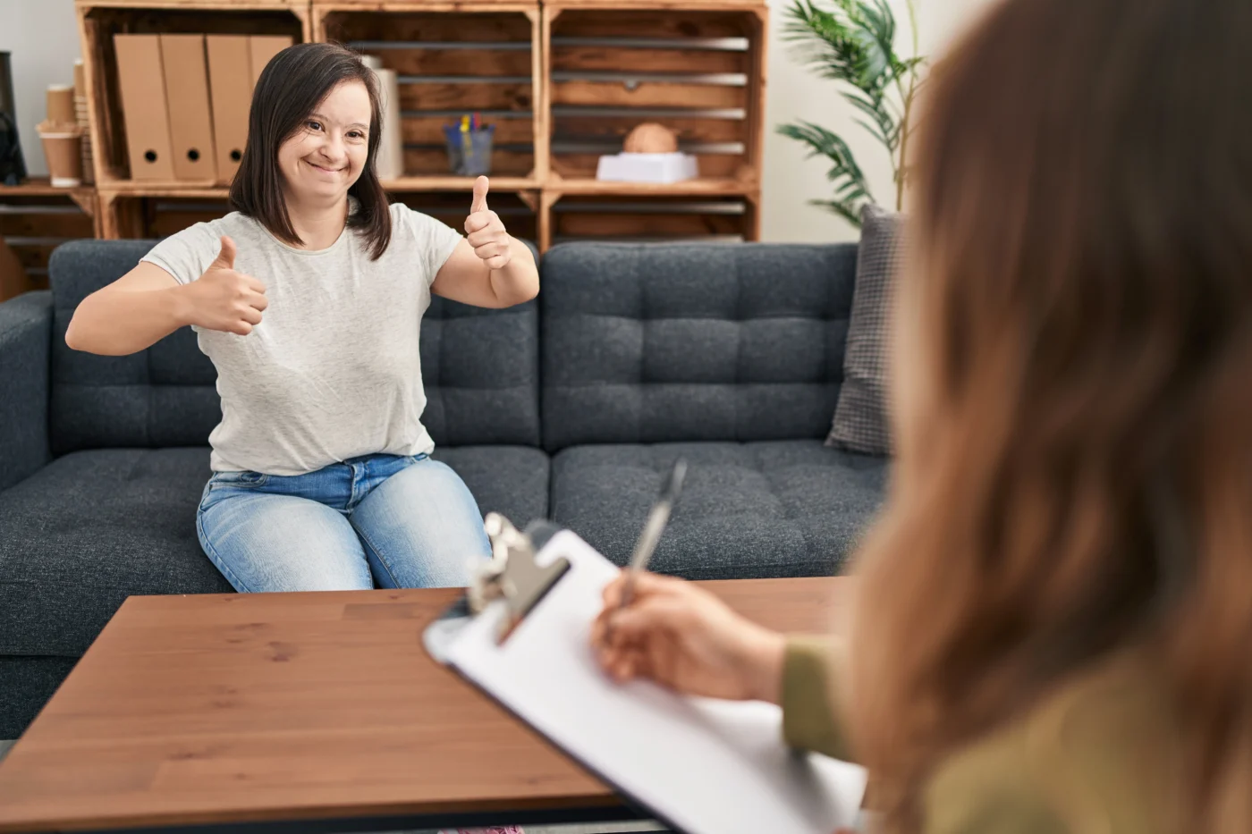 Person sitting on a couch smiling and giving a thumbs-up while speaking to another person across a table, with a clipboard in the foreground