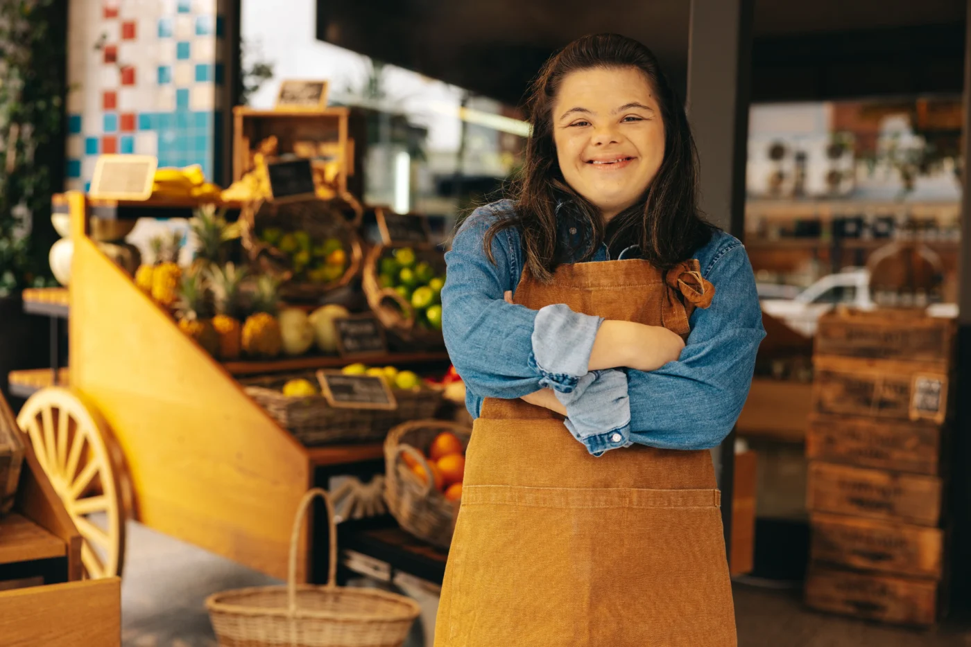 Person wearing an apron smiles with arms crossed in front of a produce display at a grocery store.