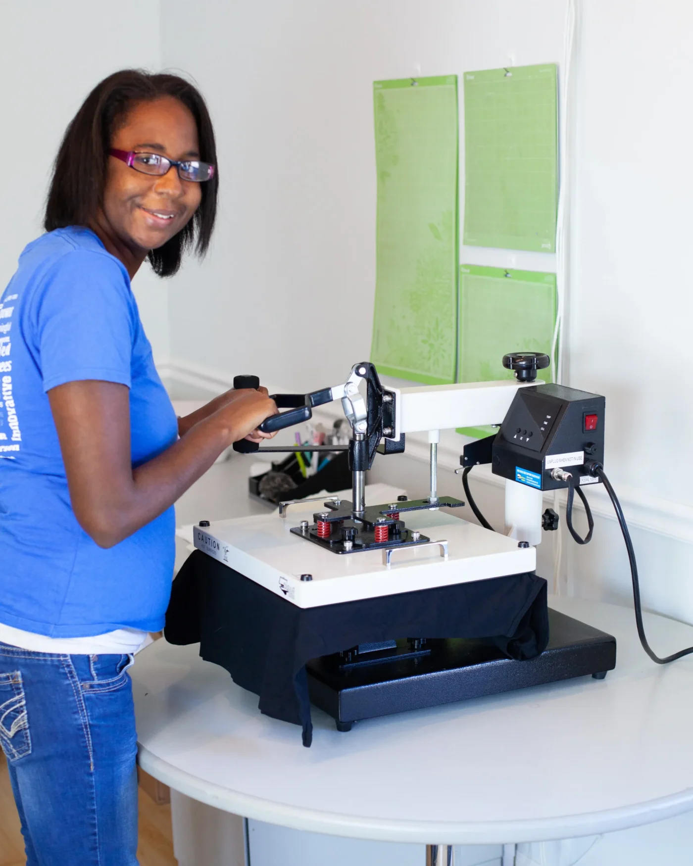 Person wearing glasses and a blue shirt smiles while operating a printing machine in a workshop.