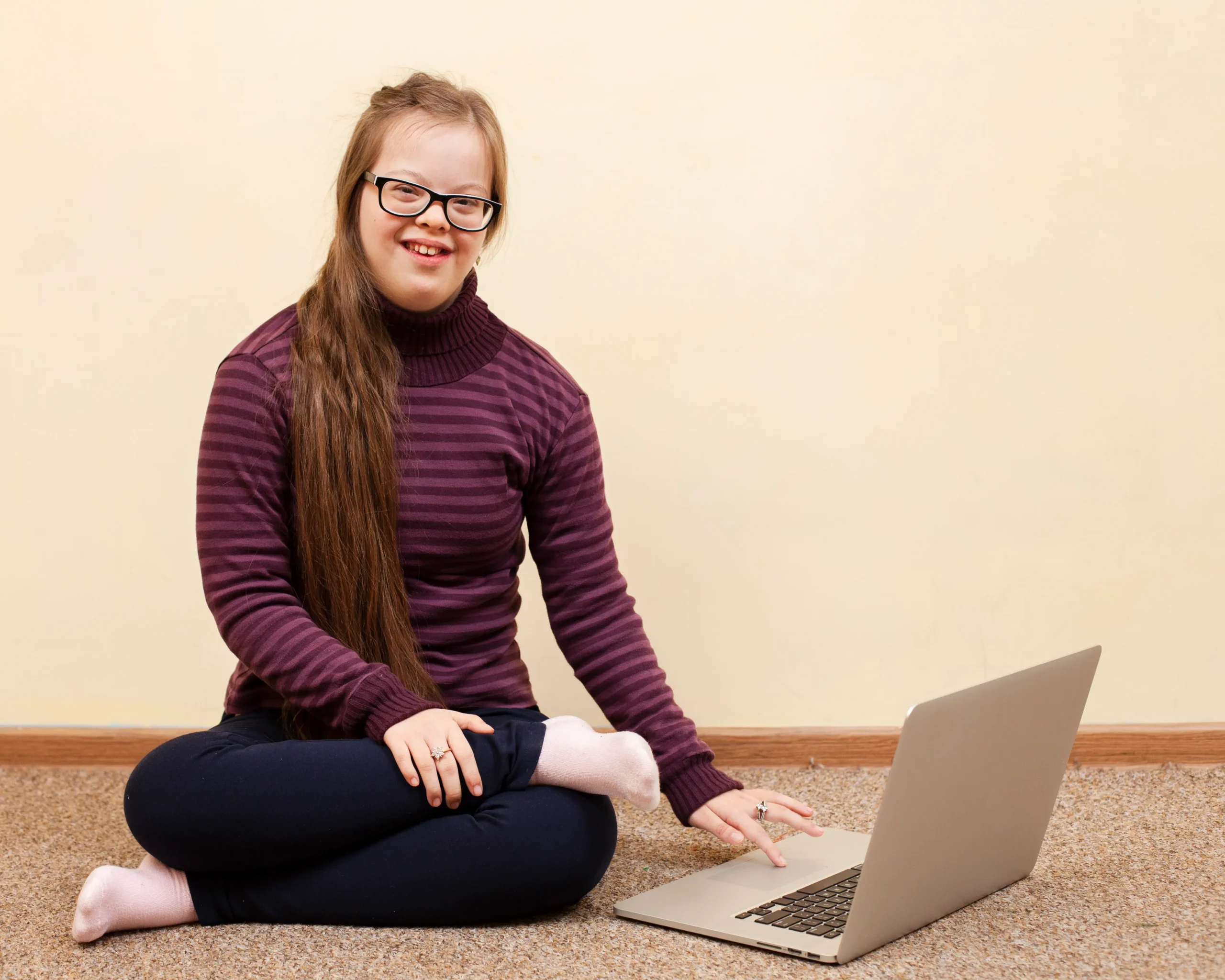 A young woman with Down syndrome sits cross-legged on a carpeted floor, smiling at the camera while using a laptop. She has long brown hair, wears glasses, and is dressed in a striped burgundy turtleneck sweater and dark pants.